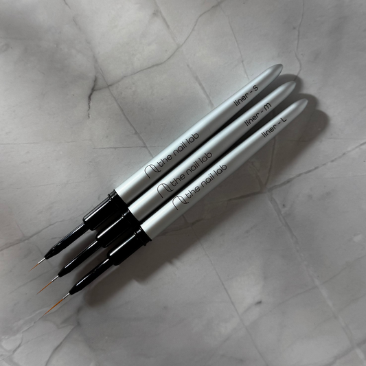 Three nail art brushes from 'The Nail Lab' on a textured gray surface